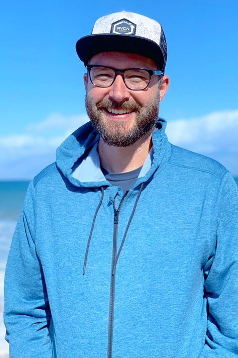 Jason Coffee in blue hoodie. Smiling at camera with sea in his background