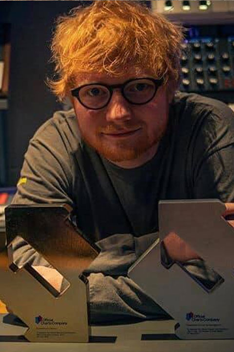Ed Sheeran wearing his glasses, and grey shirt. His golden hair are messy and his golden beard is trimmed.