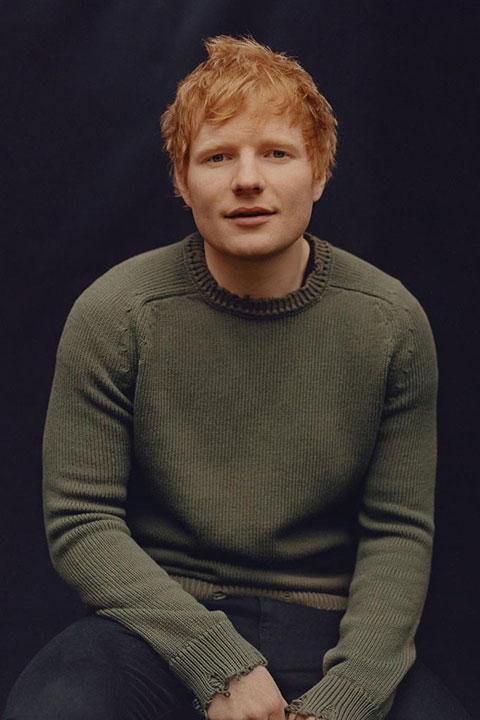 Ed Sheeran is sitting and posing for a picture in green shirt.